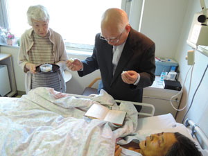 病院のベッド脇でパンを裂く牧田牧師。パンとぶどう汁のお膳を支えている牧田悦子夫人。